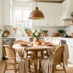 Cuisine blanche réchauffée par une table ronde en bois, des chaises tressées, des textiles beiges, des céramiques terre cuite et des plantes vertes.