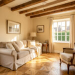 Salon lumineux d’une maison familiale rénovée avec poutres apparentes, parquet ancien, canapé en lin beige, fauteuil vintage et murs blanc cassé.